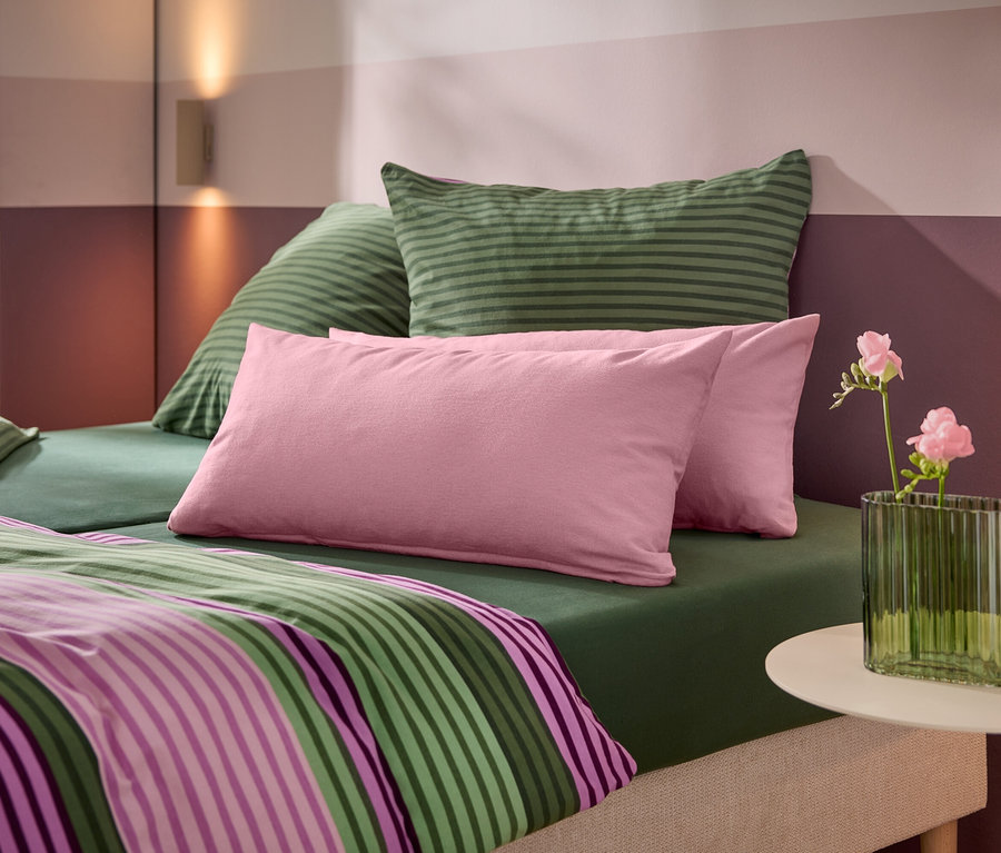 Un lit avec un drap-housse vert, des oreillers rayés verts, des oreillers roses et une couette rayée. Une table d'appoint blanche supporte un vase vert nervuré avec des fleurs roses. Le mur derrière présente des rayures horizontales roses et violettes avec deux appliques murales.