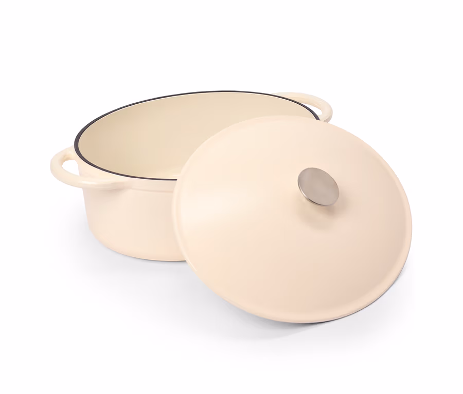 Une cocotte couleur crème avec son couvercle légèrement entrouvert à côté. La cocotte a deux poignées et un bord noir, le couvercle a un bouton argenté.