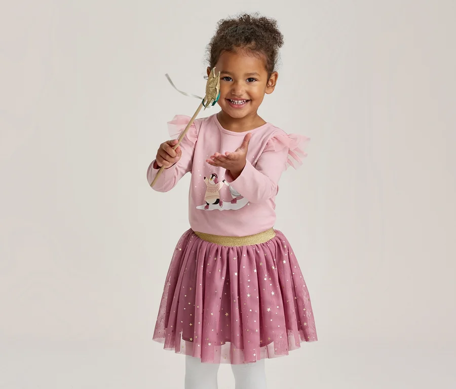 Une petite fille souriante vêtue d'un haut rose à manches longues avec motif et volants, d'une jupe en tulle rose à étoiles dorées et d'un collant blanc, tenant une baguette magique.