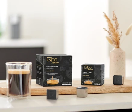 Deux boîtes noires de Qbo Caffè BABA BUDAN (27 et 8 capsules) et deux capsules de café Qbo individuelles sont présentées sur une planche en bois à côté d'un verre de café. Un vase avec de l'herbe séchée et un bol de biscuits sont également visibles.