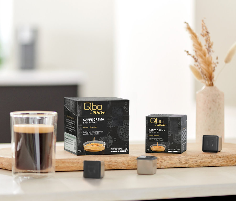 Sur une table, sur une planche en bois, se trouvent des capsules Qbo Caffè BABA BUDAN, une tasse de café et des prototypes de capsules.
