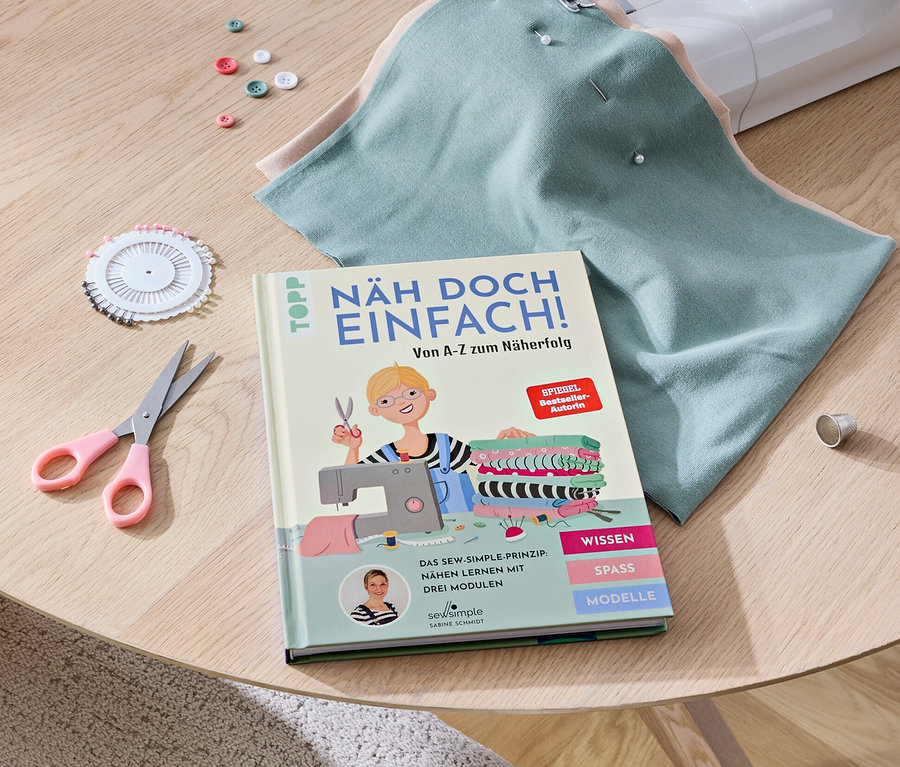 Sur une table se trouvent une machine à coudre, le livre « Näh doch einfach », des tissus, des ciseaux, des boutons et des épingles.