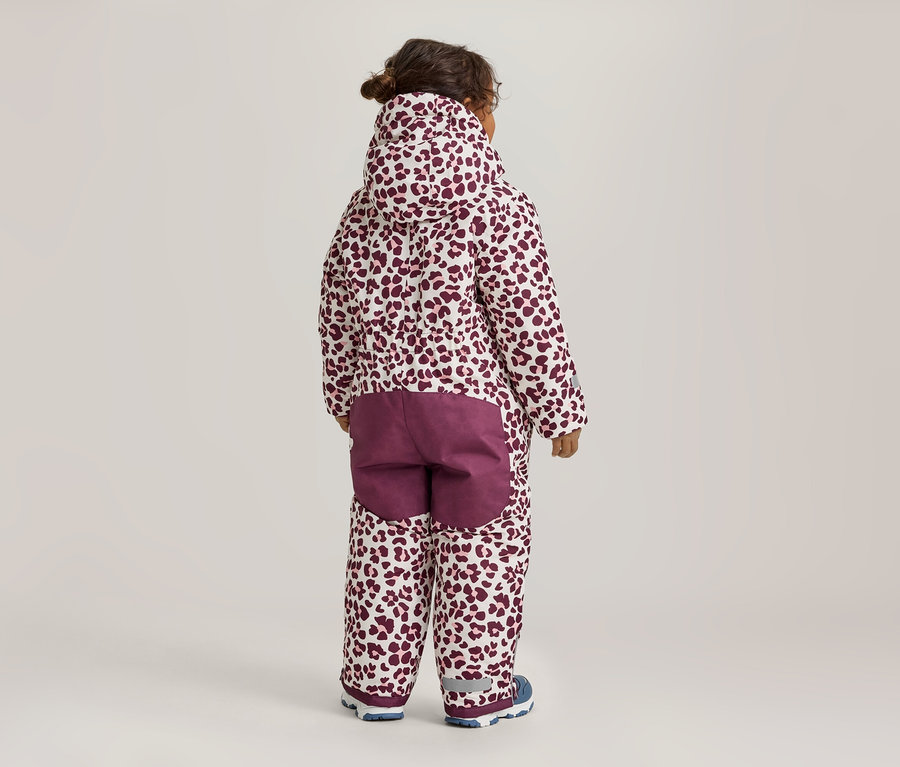 Enfant debout de dos, portant une combinaison de neige blanche à imprimé léopard rouge foncé, avec des empiècements bordeaux unis sur les fesses et des chaussures bleues et blanches.