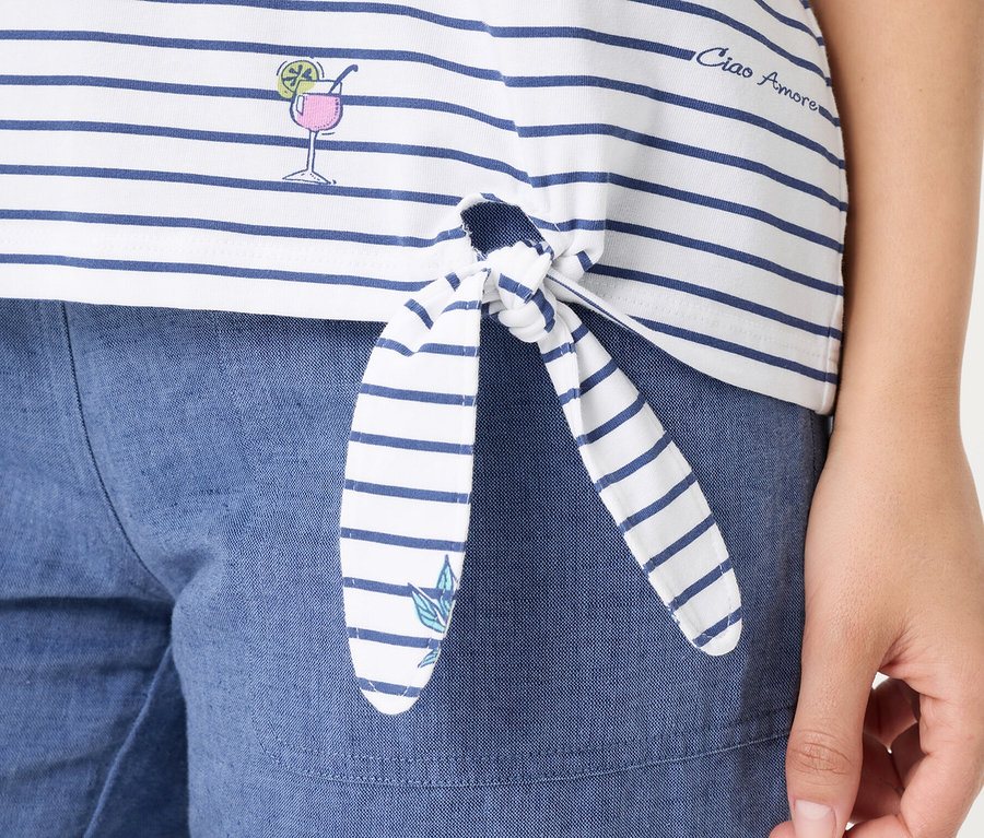 Gros plan d'une personne portant un T-shirt Olsen blanc à rayures bleues, noué sur le devant. Le T-shirt présente un verre à cocktail brodé, le texte 'Ciao Amore' et une petite feuille sur une extrémité du nœud, associé à un short bleu.