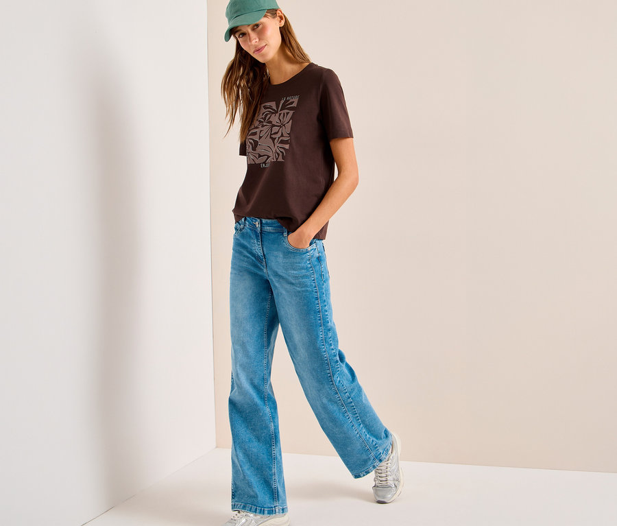 Une femme portant une casquette verte, un T-shirt à motif CECIL marron et un jean bleu, debout, la main dans la poche.
