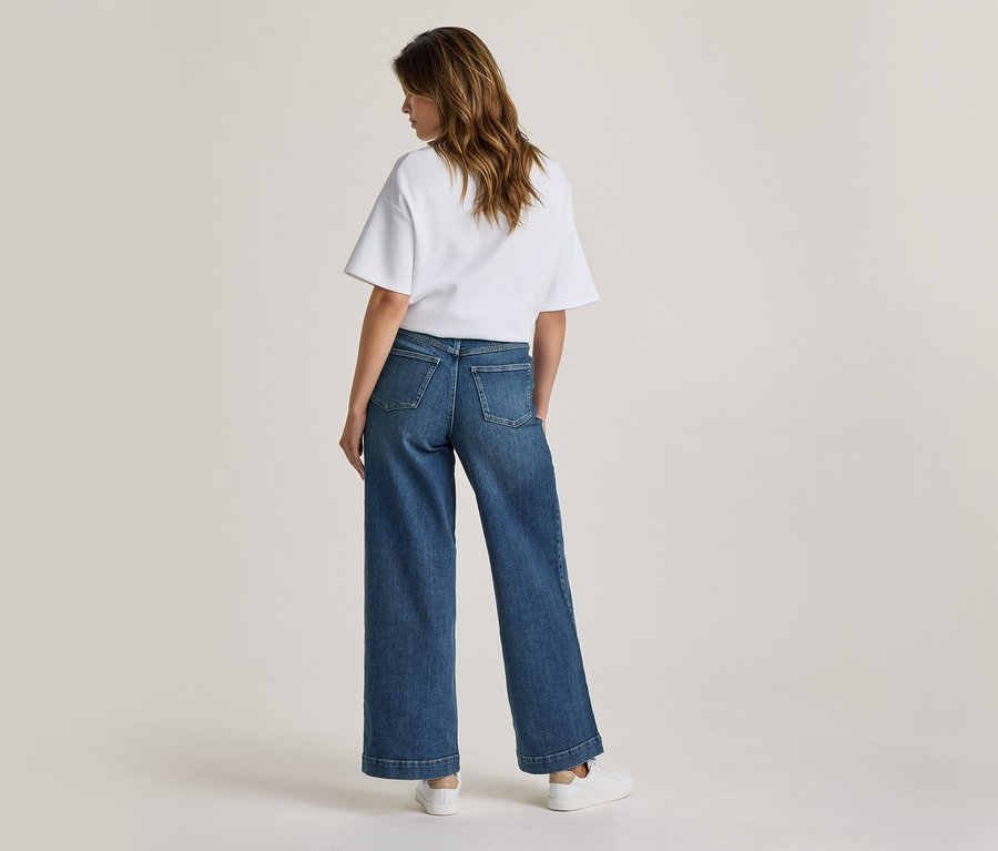 Femme debout de dos, portant un t-shirt blanc, un jean large bleu et des baskets blanches.