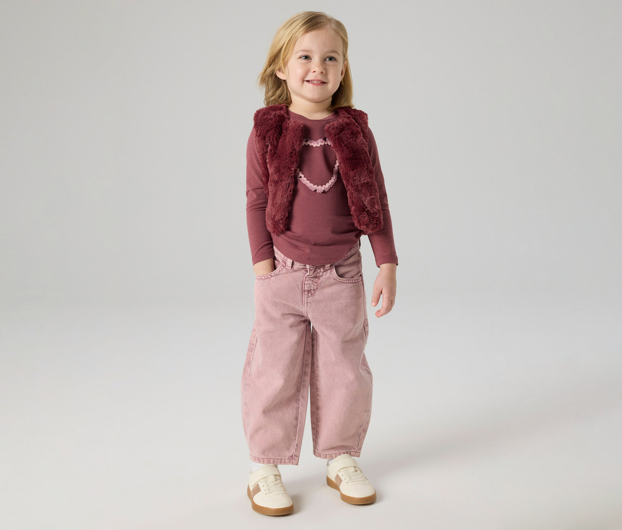 Jeune fille blonde portant un haut à manches longues couleur baie, un gilet en fausse fourrure couleur baie avec un détail cœur rose clair, un pantalon large rose avec les mains dans les poches et des baskets claires, debout et souriante.