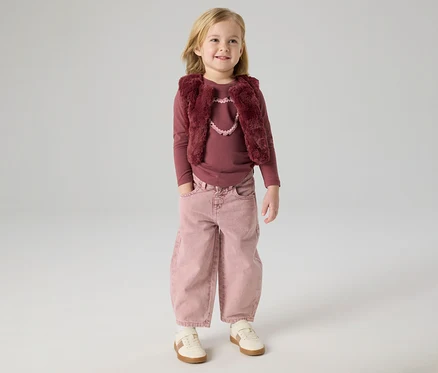 Jeune fille blonde portant un haut à manches longues couleur baie, un gilet en fausse fourrure couleur baie avec un détail cœur rose clair, un pantalon large rose avec les mains dans les poches et des baskets claires, debout et souriante.