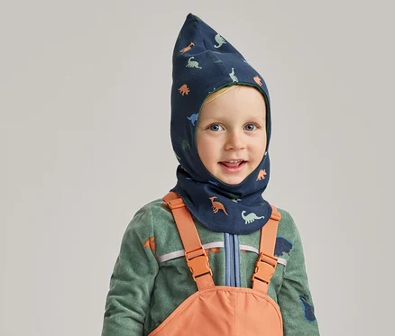 Un garçon souriant portant une cagoule bleue à motifs de dinosaures, un pull en polaire vert et une salopette orange sur fond gris.