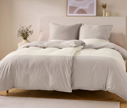 Un lit avec une parure de lit en flanelle taupe et blanc cassé. La housse de couette a une partie supérieure et inférieure taupe avec une bande centrale blanc cassé, et deux oreillers taupe reposent contre une tête de lit claire.