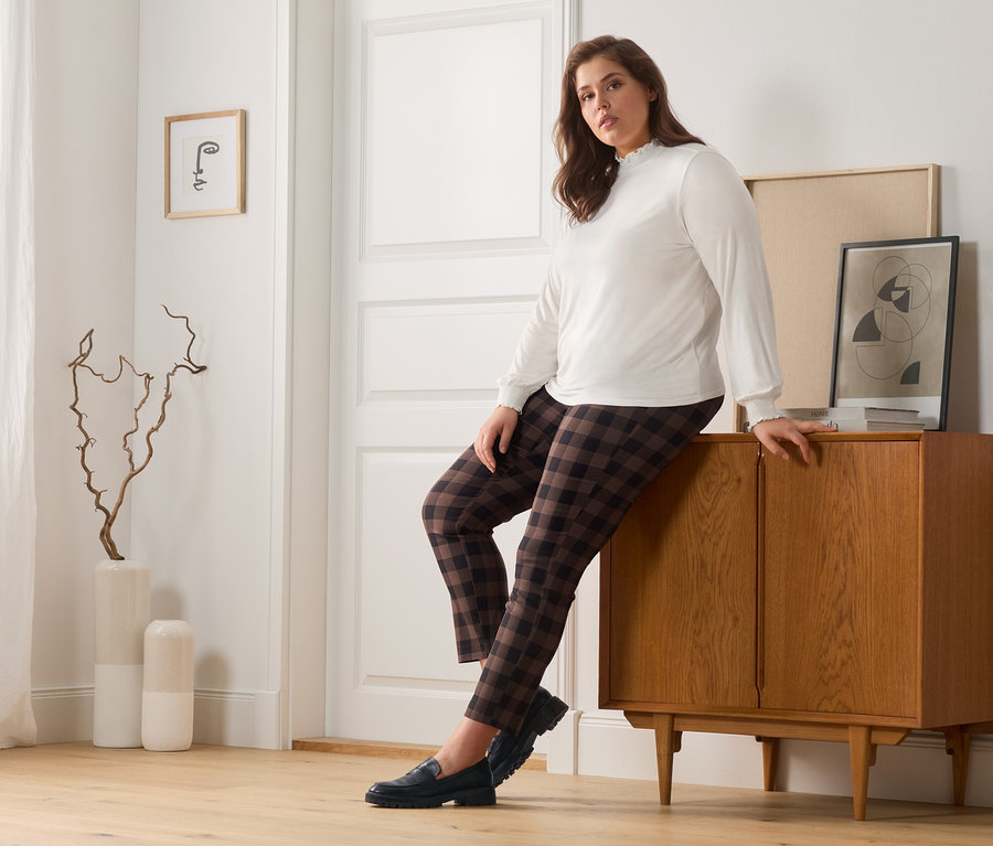 Une femme est appuyée contre une commode en bois, elle porte un pantalon stretch à carreaux, une blouse à col montant et des loafers en cuir.