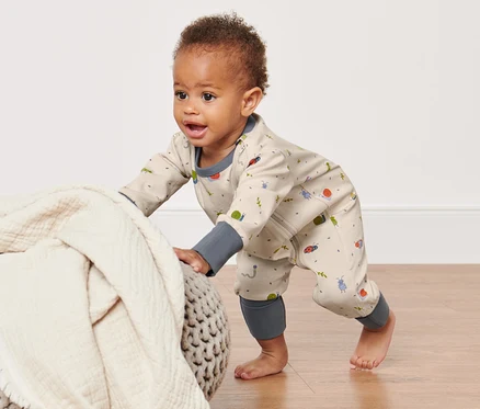 Bébé rampant en pyjama avec fermeture éclair s'appuyant sur un pouf recouvert d'une couverture.