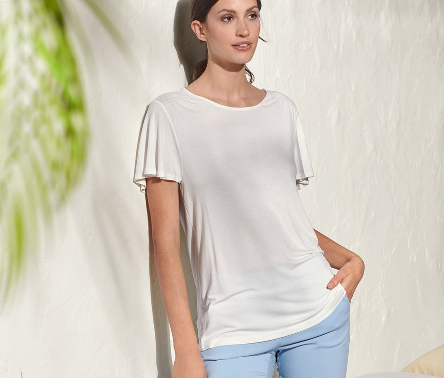 Une femme pose dans un T-shirt blanc à manches papillon et un pantalon bleu.