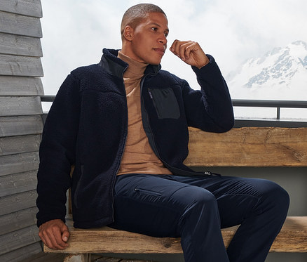 Un homme est assis sur un banc et porte un pantalon thermique, une veste en polaire et des chaussures marron.