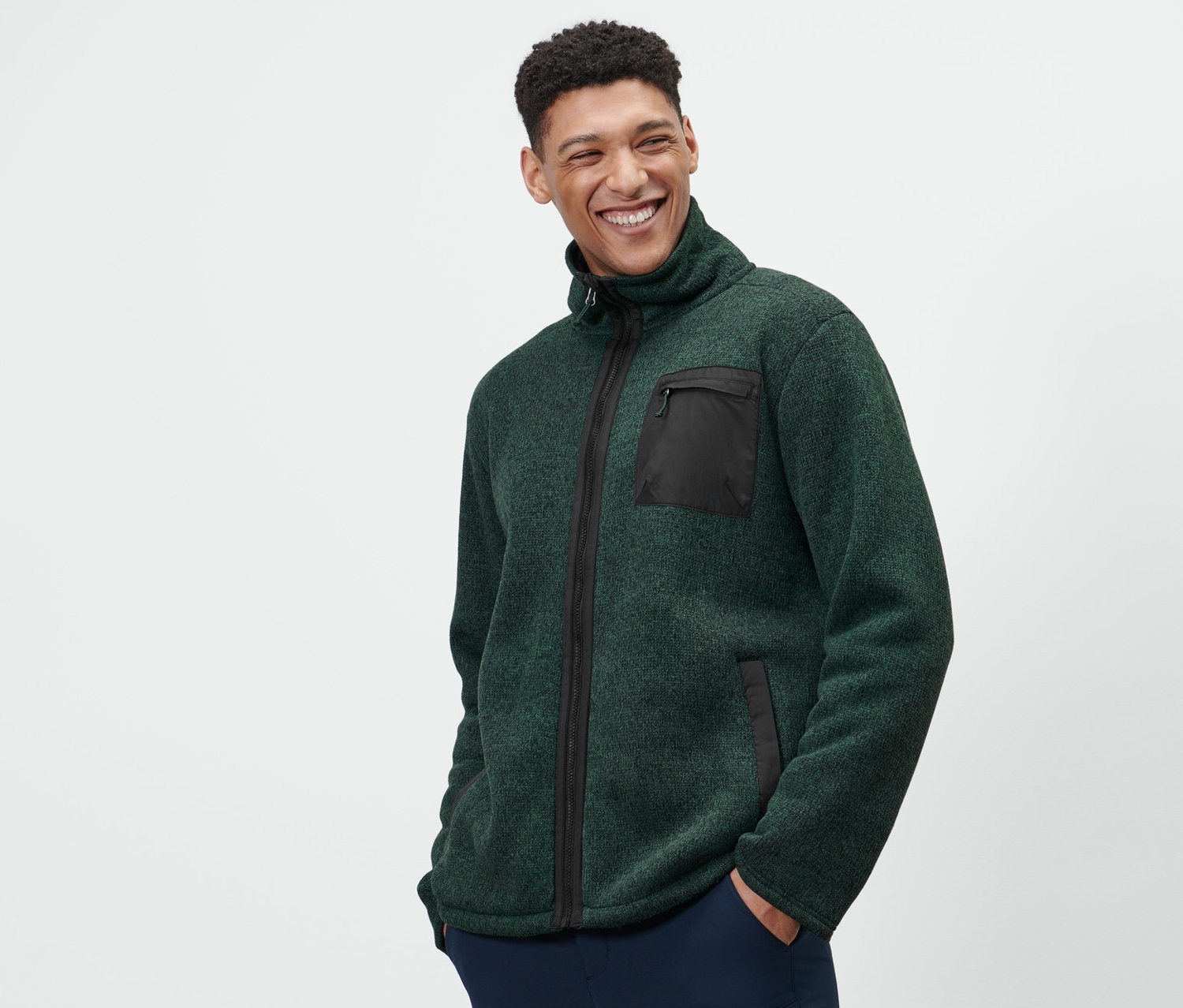 Un homme porte une jaquette en matière polaire tricotée thermique verte et un pantalon bleu marine.