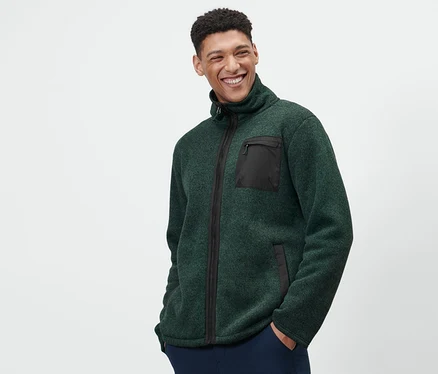 Un homme porte une jaquette en matière polaire tricotée thermique verte et un pantalon bleu marine.