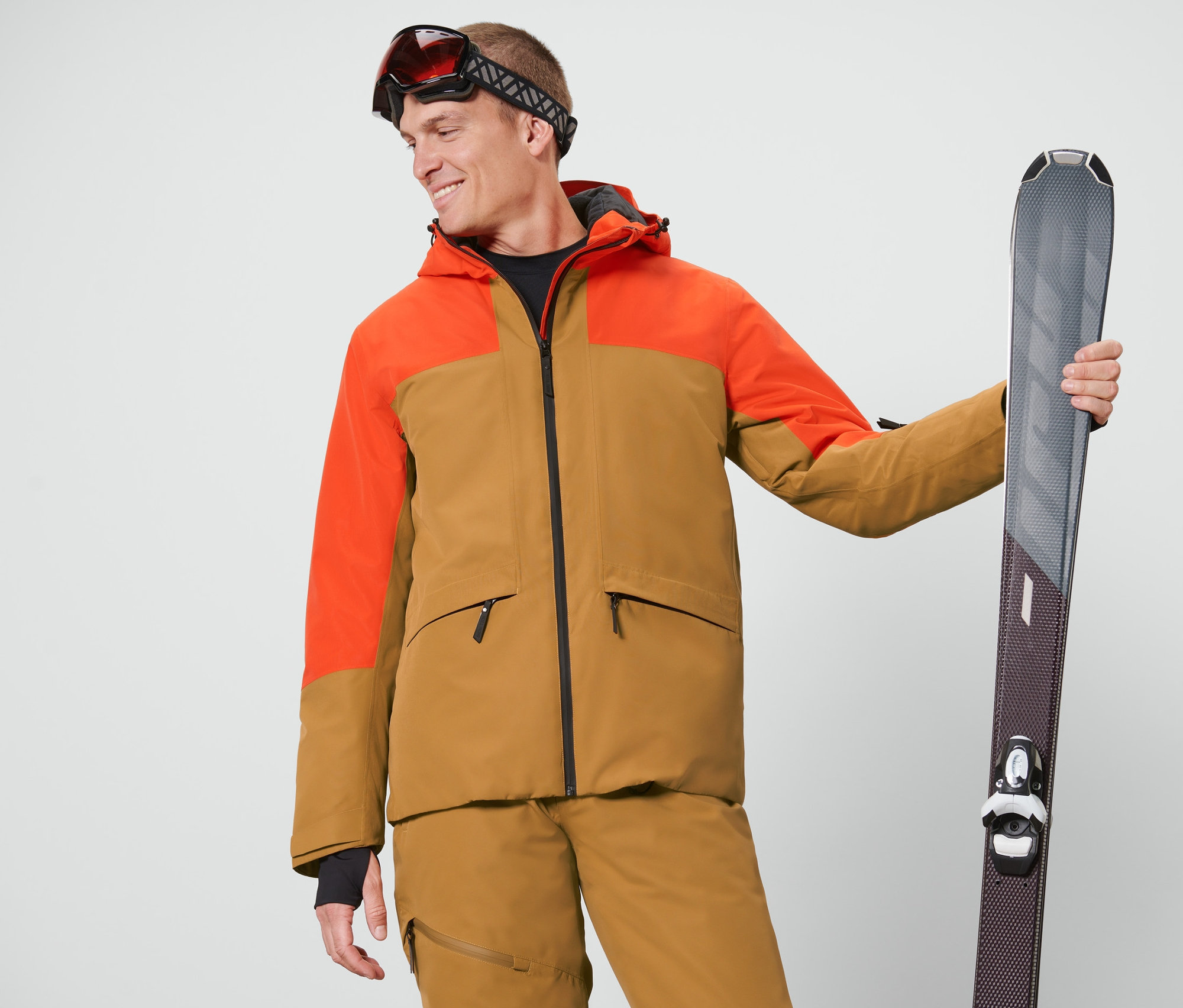 Un homme portant des lunettes de ski tient des skis et porte une veste de ski orange et marron.
