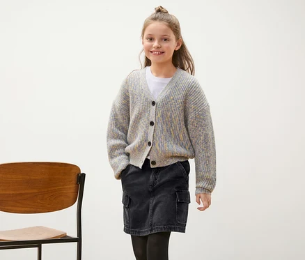 Fillette souriante posant en cardigan gris et jupe en jean noire.