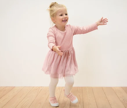 Une fillette blonde souriante en robe à côtes pour enfants pose sur un plancher en bois.
