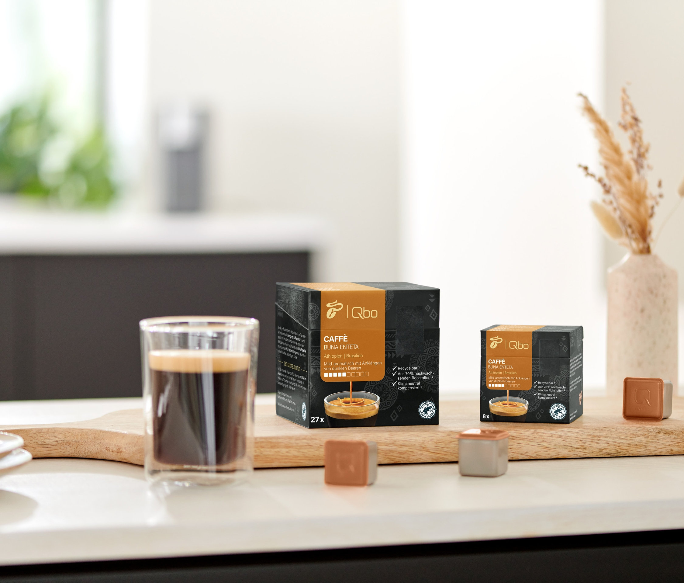 Sur une planche en bois se trouvent un verre de café et des paquets de Qbo Caffè BUNA ENTETA.