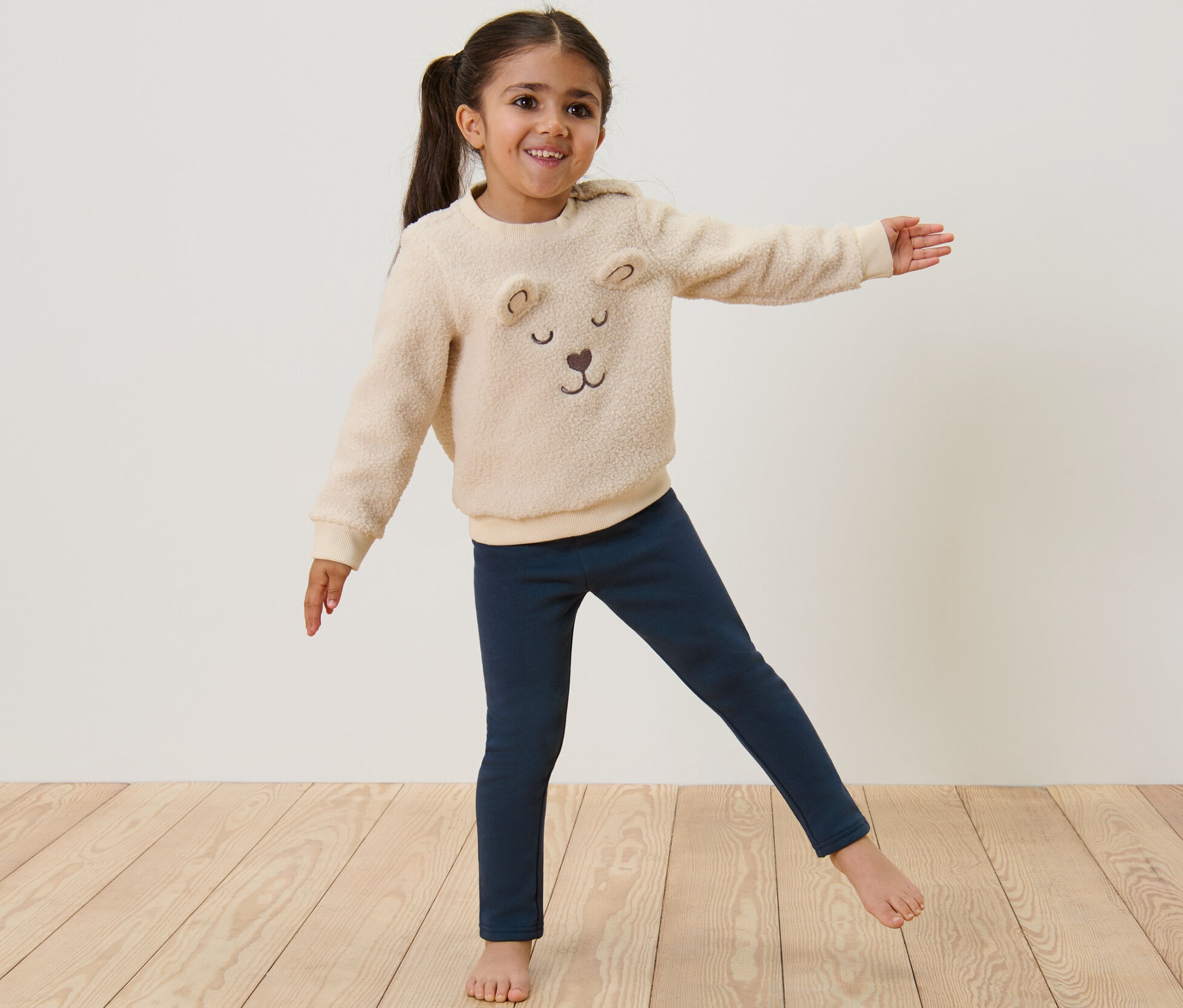 Une fille debout, les bras tendus, portant un legging thermique bleu pour enfants et un sweat-shirt ours en peluche.