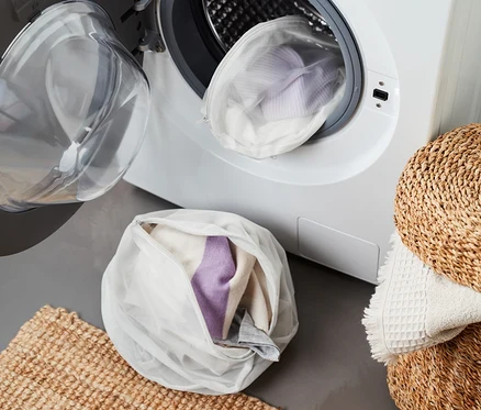 Deux filets de lavage, dont un XL, remplis de vêtements, sont posés devant un lave-linge ouvert.