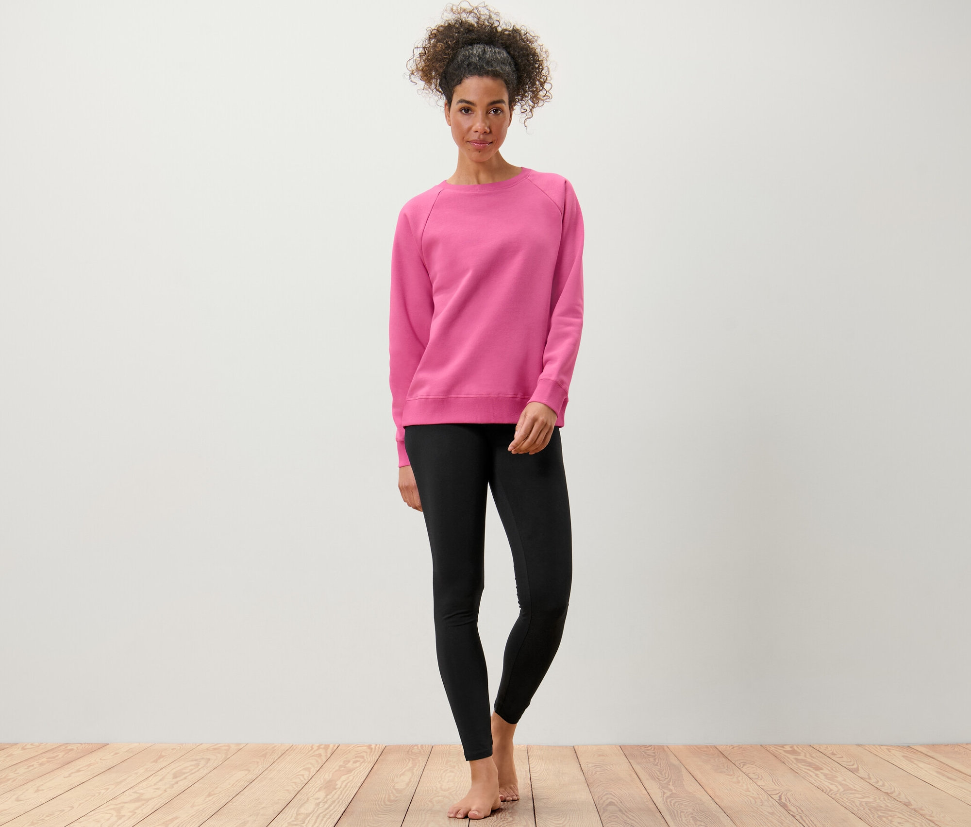 Une femme debout, pieds nus, portant un sweatshirt de yoga rose et un legging noir.