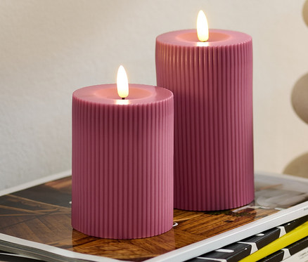 Deux bougies à LED en cire véritable, rose, sont posées sur une pile de magazines. Un vase avec de l'eucalyptus est visible en arrière-plan.