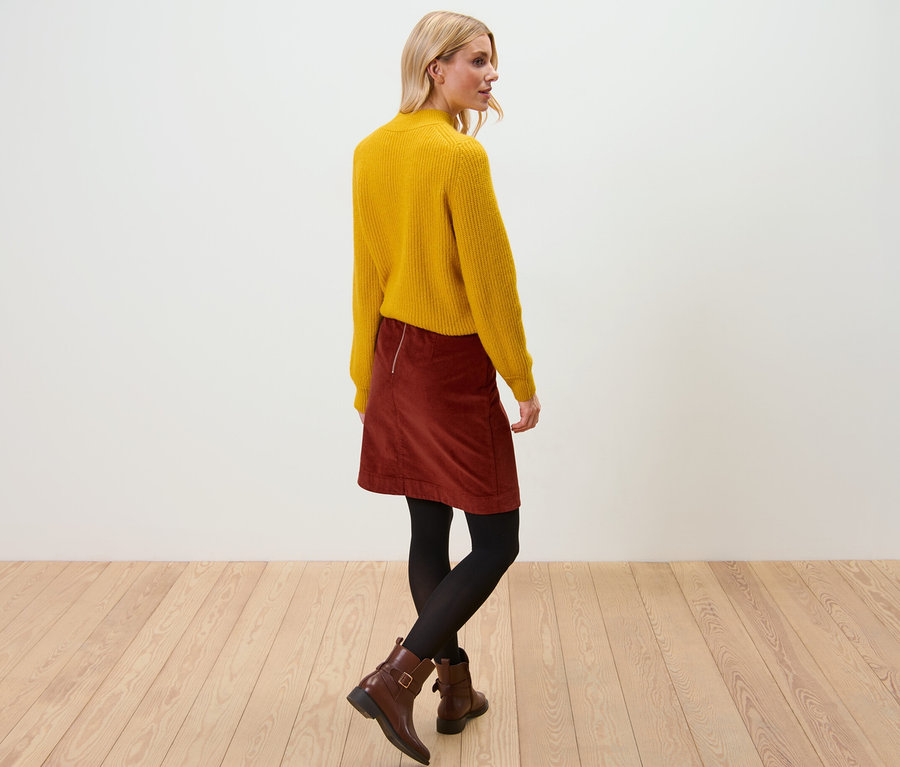 Femme blonde portant un pull jaune et une jupe en velours. Elle se tient sur un plancher en bois.