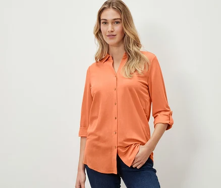 Une femme porte un chemisier en jersey piqué pêche et un jean.