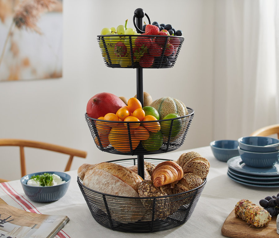 Un présentoir à étages noir à trois niveaux en métal est posé sur une table, rempli de fruits et de produits de boulangerie.