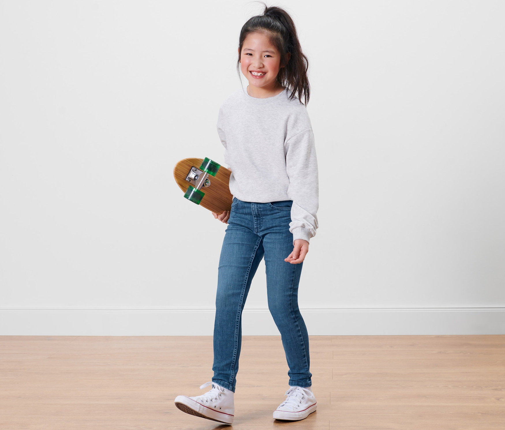 Une fillette souriante tient un skateboard et porte un jean taille haute bleu pour enfant – Coupe « Lotta ».