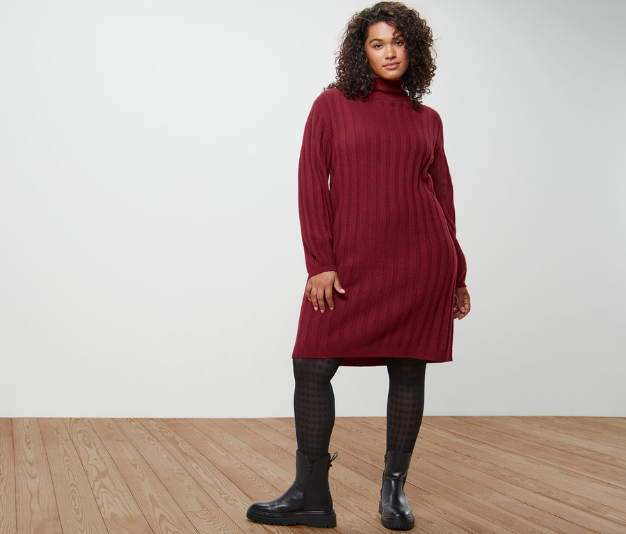 Femme posant en robe en maille côtelée avec col montant, collants fins à motifs et bottines en cuir.