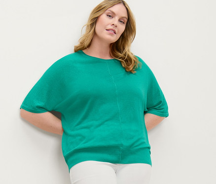 Une femme pose contre un mur, portant un haut à maille fine avec du lin et un tregging 7/8 à broderie ajourée.