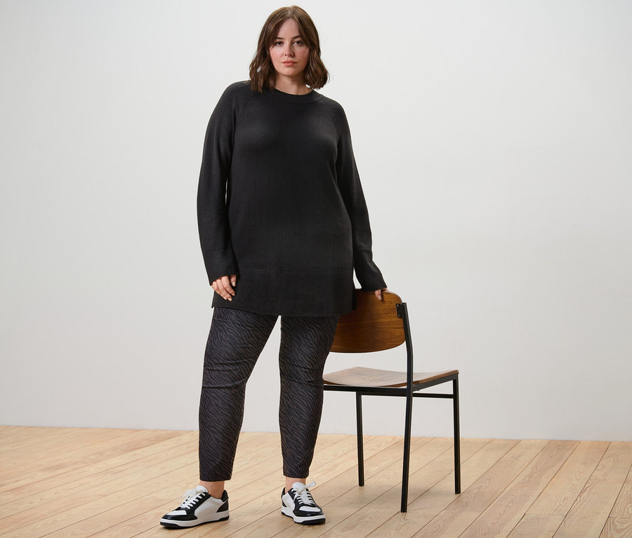 Une femme pose en long pull en maille noir et pantalon imprimé en bengaline à côté d'une chaise.