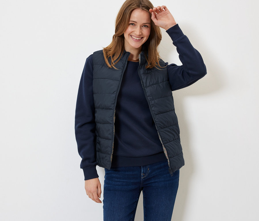 Une femme souriante pose dans un gilet matelassé bleu marine, un pull et un jean.