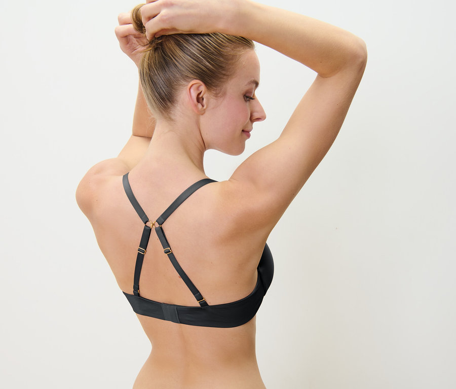 Une femme blonde attache ses cheveux en chignon, portant un soutien-gorge noir à bretelles décoratives.