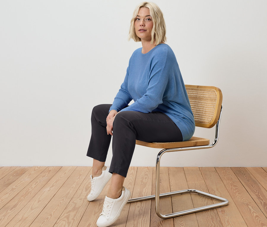 Femme blonde assise sur une chaise portant un pantalon stretch et un pull en maille fine bleu.