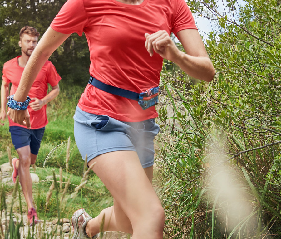 Une femme portant un T-shirt fonctionnel corail et un legging de sport 3/4 bleu foncé court sur un chemin rocailleux. À l'arrière-plan, un homme court en short fonctionnel et en T-shirt de sport corail.