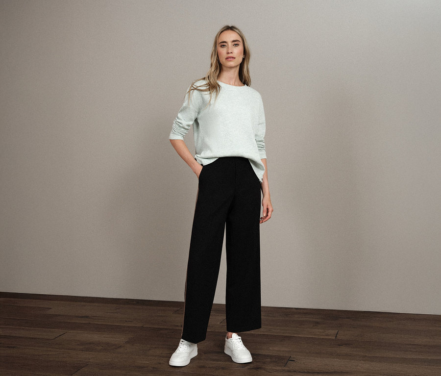 Femme debout, la main dans la poche, portant un maillot avec structure Street One, un pantalon noir et des baskets blanches.