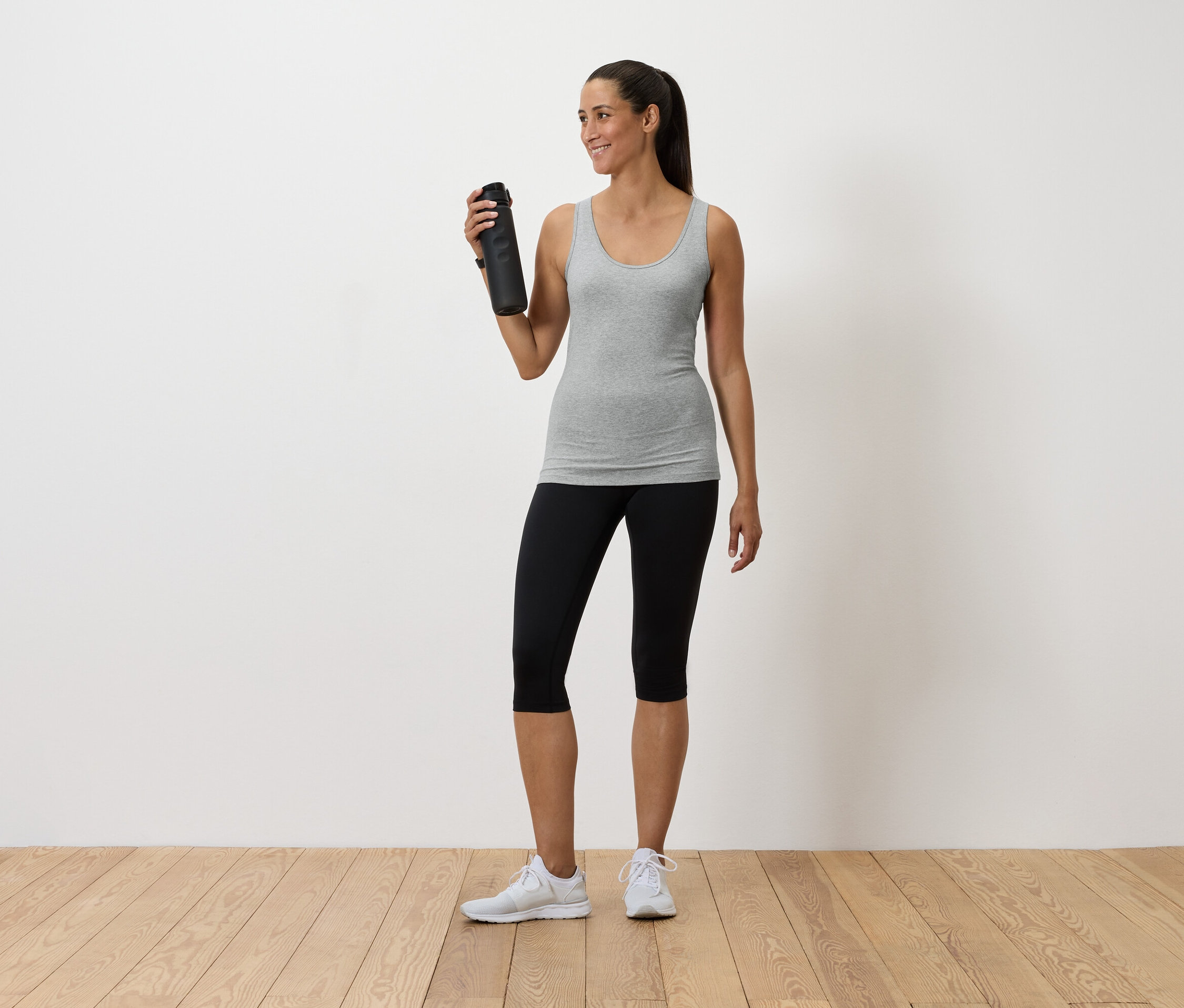 Femme tenant une bouteille d'eau, portant un débardeur gris, un legging noir et des baskets blanches.