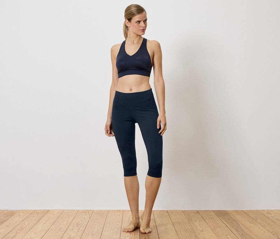 Une femme en soutien-gorge de sport bleu marine et leggings debout sur un plancher de bois.