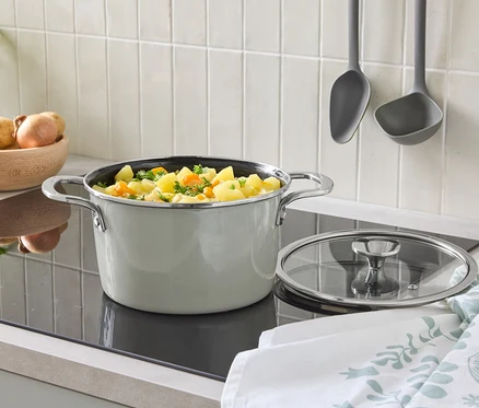 Sur une plaque de cuisson se trouve une casserole avec un couvercle et des légumes. À l'arrière-plan, il y a un bol de pommes de terre et des louches suspendues.