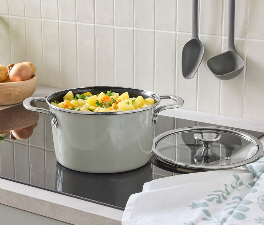 Sur une plaque de cuisson se trouve une casserole avec un couvercle et des légumes. À l'arrière-plan, il y a un bol de pommes de terre et des louches suspendues.
