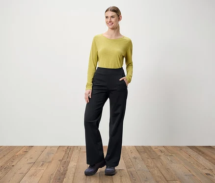 Une femme se tient sur un plancher en bois. Elle porte un chemisier vert à manches longues et un pantalon noir.