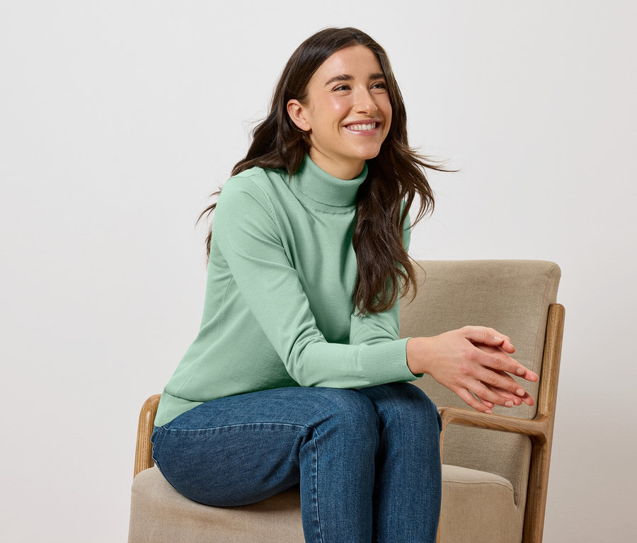 Une femme souriante est assise sur une chaise beige, portant un pull à col roulé vert et un jean.