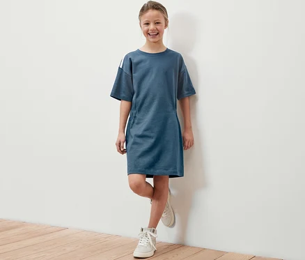 Fillette souriante posant en robe en heavy jersey bleue pour enfant avec des chaussures blanches.