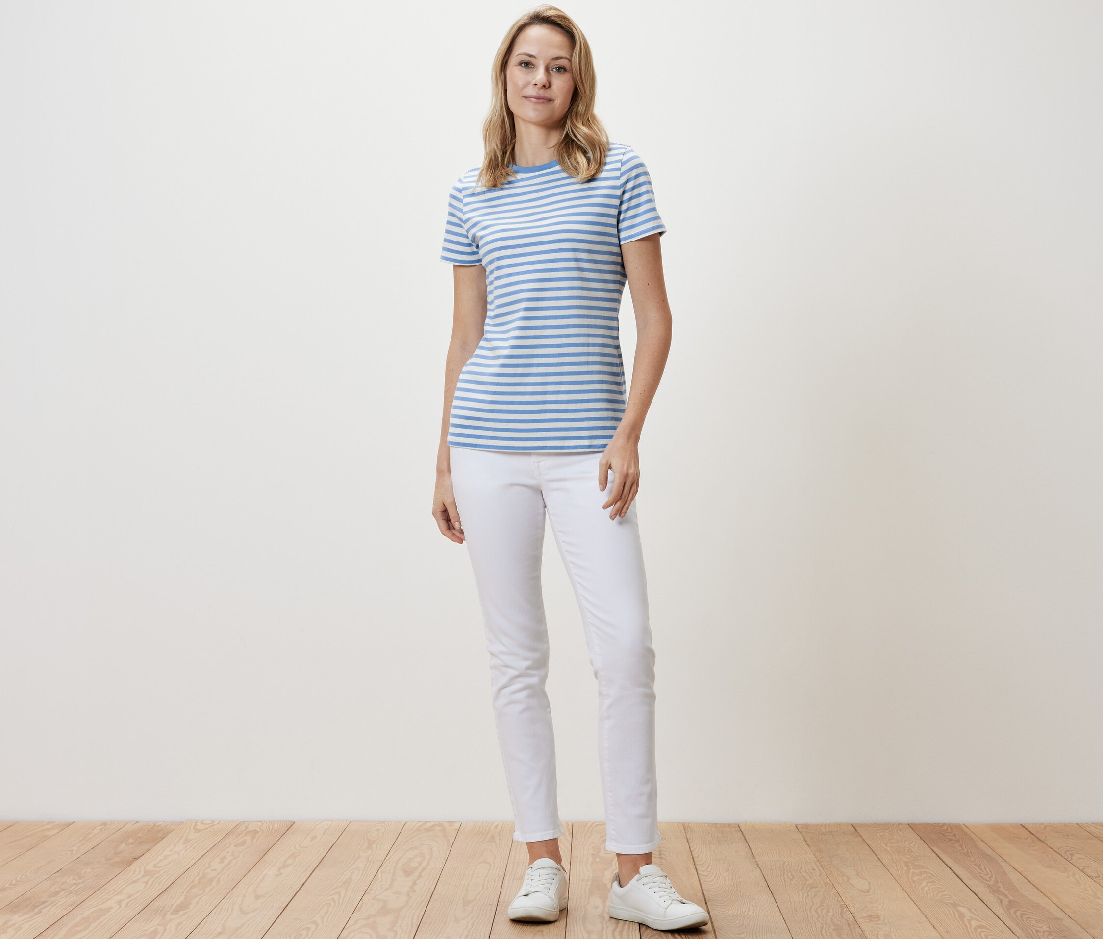 Une femme debout porte un t-shirt basique bleu clair-blanc à rayures, un pantalon blanc et des baskets blanches.