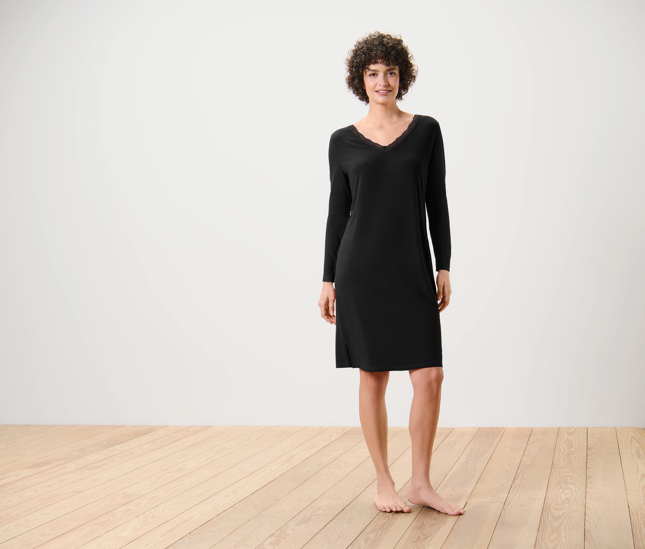 Une femme se tient pieds nus sur un plancher en bois et porte une robe noire à manches longues.