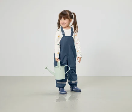 Fillette souriante avec des couettes tenant un arrosoir vert clair, vêtue d'un pantalon bleu et de bottes de pluie.